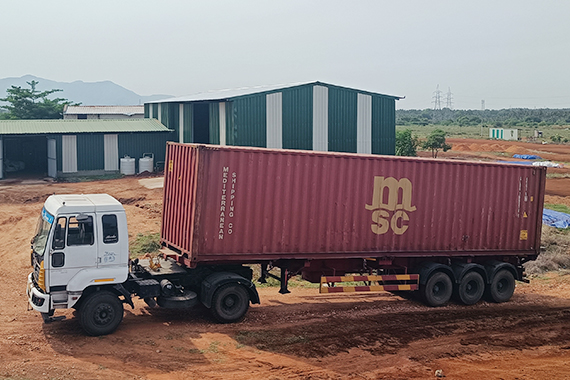 A large truck with a red MSC shipping container is parked on a dirt road. Green-roofed industrial buildings are in the background, with distant mountains and power lines visible under a cloudy sky.